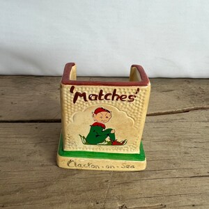 May include: A vintage ceramic match holder, beige with a textured finish, and a green base. It features a cartoon elf illustration and the word 'Matches' in red. The base is inscribed with 'Clacton-on-Sea'.