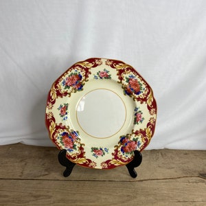 May include: A decorative plate with a cream-colored center and a burgundy rim. The rim features floral patterns in pink, blue, and green. The plate is displayed on a black stand against a white backdrop.