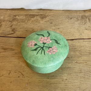 May include: A round, light green trinket box with a lid. The lid is decorated with a cluster of pink flowers and green stems. The box is sitting on a wooden surface.