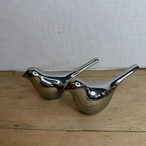 Könnte beinhalten: Zwei polierte silberne Vogelfiguren. Die Vögel haben eine glatte, reflektierende Oberfläche mit einem abgerundeten Körper und einem angehobenen Schwanz. Sie stehen auf einer hellbraunen Holzoberfläche vor einem schlichten weißen Hintergrund.