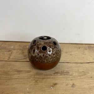 Pode incluir: Um vaso esférico de cerâmica marrom com uma parte superior salpicada e vidrada e uma base lisa marrom escuro. A parte superior apresenta várias aberturas circulares. O vaso está sobre uma superfície de madeira.