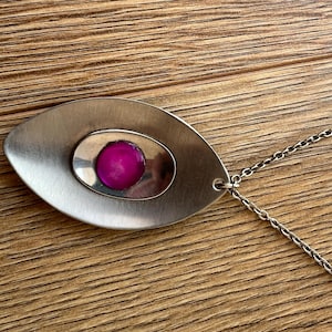 Könnte beinhalten: Eine silberfarbene Anhängerkette mit einem großen, oval geformten Anhänger. Der Anhänger hat einen zentralen, ovalen Einsatz mit einem leuchtend magenta farbenen Stein. Eine filigrane Kette vervollständigt die Halskette.