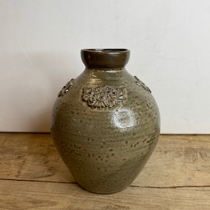 May include: A handmade, olive-green ceramic vase with a rounded body and a dark brown rim. The vase features embossed floral designs around the upper portion. The surface has a speckled texture, giving it a rustic appearance. The vase sits on a wooden surface.