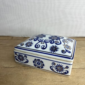 Puede incluir: Caja de baratijas de cerámica blanca con diseño floral azul y ribete dorado. La caja rectangular tiene una tapa ligeramente curvada. El patrón floral incluye flores estilizadas y remolinos. Colocada sobre una superficie de madera.