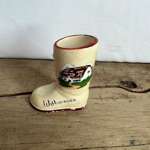 May include: A vintage ceramic boot-shaped souvenir. The boot is off-white with a painted scene of a house and the word "Withernsea" in red. The rim and base of the boot are also red.