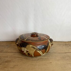 May include: A ceramic casserole dish with a lid. The dish is mainly reddish-brown with black and tan abstract designs. It has two handles and a knob on the lid. The dish is on a wooden surface.