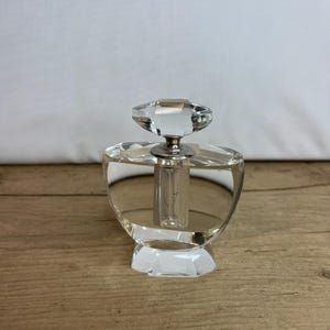 Könnte beinhalten: Eine durchsichtige Glas-Parfümflasche mit einem facettierten Stopfen. Die Flasche hat einen runden Körper und einen facettierten Boden.