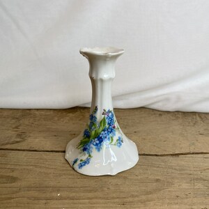 May include: White porcelain candlestick holder with a flared base and a slender, fluted stem. The base is decorated with blue floral sprigs and green leaves. The top has a scalloped edge.