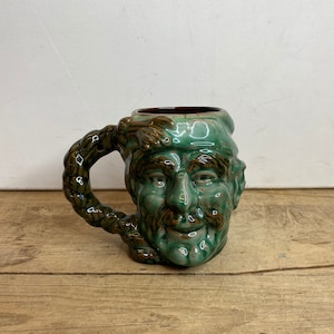 Peut inclure: Une tasse en céramique verte avec un visage sculpté et une anse tressée. Le visage a des détails bruns et une barbe. La tasse est posée sur une surface en bois.