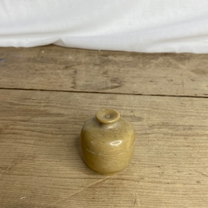 May include: A small, round, beige stone container with a lid. The container has a smooth, polished surface and sits on a wooden surface. The lid is a small, circular shape with a raised center.