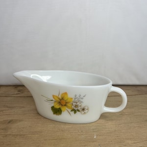May include: White ceramic gravy boat with a handle and a floral design featuring yellow and white flowers. The boat has a curved spout and a rounded body, ideal for serving sauces and gravies.