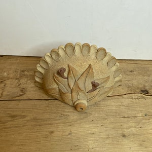 May include: A tan ceramic wall pocket with a floral design. The pocket has a scalloped edge and features stylized leaves and two small, brown, bud-like accents. It is set against a light wooden surface.