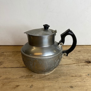 Può includere: Teiera vintage in metallo argentato con manico e coperchio neri. La teiera presenta un design ornamentale in rilievo attorno alla base e un beccuccio. Il manico è curvo e il coperchio ha un pomello nero.