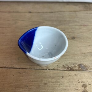 Può includere: Una piccola ciotola rotonda in ceramica con interno bianco e una sezione esterna dipinta di blu. La ciotola è appoggiata su una superficie di legno, mostrando il suo design semplice ma elegante. La vernice blu è di un colore intenso e saturo.