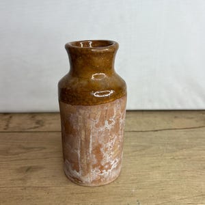 May include: A small, rustic brown ceramic vase with a cylindrical body and a slightly flared neck. The upper portion is a darker, glossy brown, while the lower half has a lighter, textured appearance. The vase is likely handmade and has a vintage aesthetic.