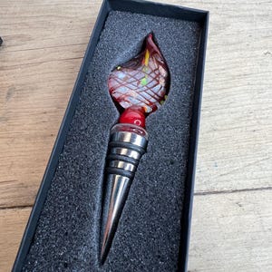 Può includere: Un tappo per bottiglia in vetro rosso e giallo a forma di fiamma, con una base in metallo argentato. Il tappo è in una scatola nera con una fodera in schiuma grigia.