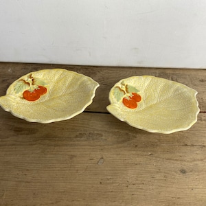 Puede incluir: Dos platos de cerámica con forma de hoja. Los platos son de color amarillo claro con una superficie texturizada. Cada plato tiene un diseño de dos tomates naranjas con hojas verdes. Los platos están sobre una superficie de madera.