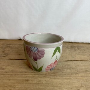 May include: A small, white ceramic plant pot with a hand-painted floral design. The flowers are pink and purple with green leaves. The rim is a light purple colour. The pot sits on a wooden surface.