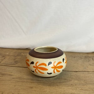 May include: A round ceramic vase with a cream-colored base, a dark brown band at the top, and orange floral designs. The vase has a small opening and is set on a wooden surface.