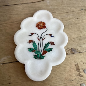 May include: White marble coaster with a floral inlay design. The coaster features a flower with orange petals, green leaves, and accents of blue and brown. The coaster has a unique, scalloped edge.