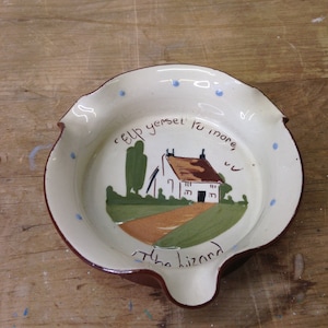 May include: A ceramic ashtray with a cream-colored interior and a brown rim. The ashtray features a hand-painted scene of a house, trees, and fields, with the text "elp yersel tu more, the lizard" around the edge.