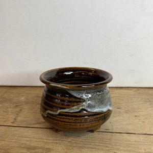 May include: A small, handcrafted ceramic pot with a rounded shape and three small feet. The pot features a dark brown glaze with horizontal bands of lighter brown and white, creating a textured appearance. The rim is a darker, glossy brown.