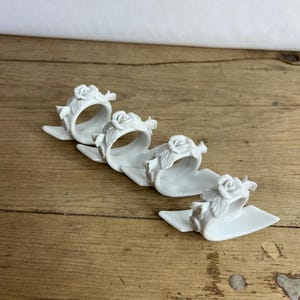 Könnte beinhalten: Set aus vier weißen Porzellan-Serviettenringen mit einem erhabenen Rosenmuster. Jeder Ring ist blattförmig und hat ein zartes florales Detail.