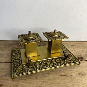 May include: A vintage brass inkwell set with two inkwells and a pen holder. The inkwells have ornate lids and the base is decorated with a floral design.