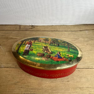 May include: Oval red tin with a gold rim and a painted pastoral scene on the lid. The scene depicts a picnic with figures, a dog, and a cart on a green field. The tin's side has a decorative pattern.