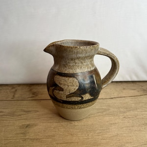May include: A small, handmade ceramic pitcher with a handle and spout. The pitcher features a beige base, a brown and black patterned design, and a textured, speckled finish. The pitcher is displayed on a wooden surface.