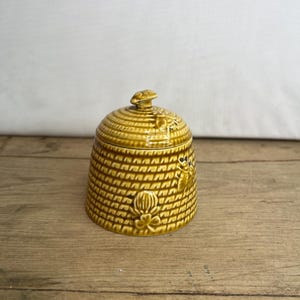 Könnte beinhalten: Keramik-Honigtopf mit Deckel, im Bienenwaben-Design. Der Topf hat eine warme gelbbraune Farbe mit strukturierter Oberfläche und ist mit Bienen- und Blumenmotiven verziert. Der Deckel hat einen kleinen Knopf.