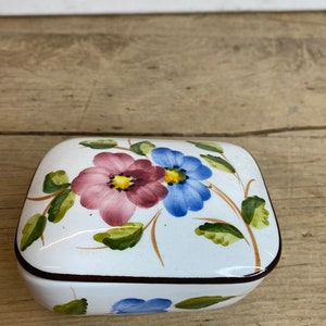 May include: A white, square trinket box with a floral design. The lid features two large flowers, one pink and one blue, with green leaves and brown stems. The box has a dark brown trim.