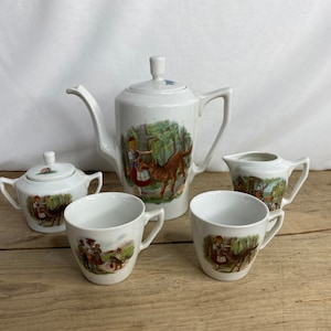 May include: A white porcelain tea set comprising a teapot, a lidded sugar bowl, a creamer, and two cups. Each item is decorated with colourful illustrations of a girl in a red skirt and a brown dog in a forest setting.