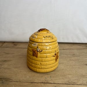 May include: A yellow ceramic beehive-shaped jar with a lid. The jar has a textured surface with horizontal lines and is decorated with brown bees. The word "Whitby" is written on the lid.