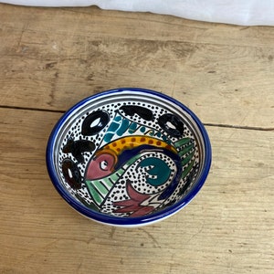 Peut inclure: Un petit bol en céramique blanche avec un bord bleu. Le bol est décoré d'un motif abstrait coloré avec des points noirs, des tourbillons verts, bleus, jaunes et roses.