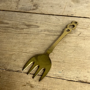 May include: A vintage brass fork with three tines and a decorative handle featuring three connected circles. The fork is set against a weathered wooden surface, showcasing its aged appearance and unique design.