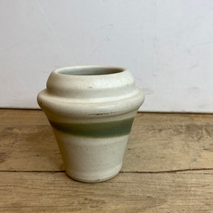 May include: A small, off-white ceramic vase with a green band around the middle. The vase has a unique, rounded shape with a flared top and a slightly textured surface. The vase sits on a wooden surface.