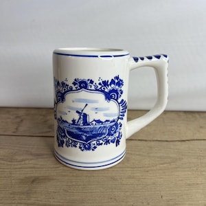 Könnte beinhalten: Weißer Keramikbecher mit blau-weißem Windmühlen-Design. Der Becher hat eine rechteckige Form und einen mit blauen Punkten verzierten Griff. Das Design zeigt eine Windmühle, Wasser und Häuser, umgeben von floralen Mustern.