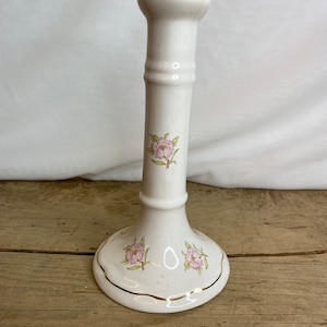 Puede incluir: Un candelero de cerámica blanca con base acampanada y tallo cilíndrico y delgado. El candelero está decorado con diseños florales rosas y ribetes dorados. El candelero mide aproximadamente 15 cm de altura.