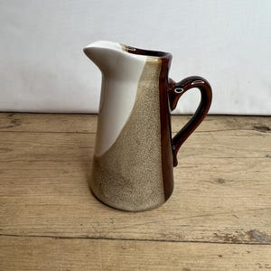 Könnte beinhalten: Eine braune, weiße und beige Keramikkanne mit braunem Henkel. Die Kanne hat eine strukturierte Oberfläche und einen leicht ausgestellten Rand.