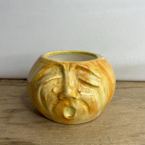 May include: A ceramic bowl with a face design. The bowl is round with a yellow and orange color scheme. The face has closed eyes and an open mouth. The interior of the bowl is white with a green rim. The bowl is sitting on a wooden surface.