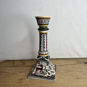 May include: A white ceramic candlestick with a square base and a cylindrical column. The base and column are decorated with colourful floral and geometric patterns. The top of the candlestick features a circular opening for a candle. The base features a painted image of a deer in a forest setting.