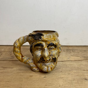 Peut inclure: Une tasse en céramique avec un visage sculpté. La tasse présente un visage détaillé avec une moustache et une barbe, et une anse torsadée. Les couleurs sont principalement marron, beige et noir. La tasse est posée sur une surface en bois.