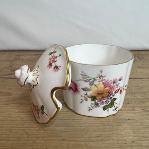 Puede incluir: Un recipiente de porcelana blanca con tapa, con diseños florales en rosa, amarillo y morado. Los bordes están adornados con oro. La tapa está parcialmente abierta, revelando el interior.