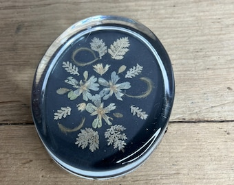 May include: Oval-shaped paperweight with a clear, rounded exterior. The interior features a dark blue background with a floral arrangement of dried flowers and leaves. The paperweight is resting on a wooden surface.