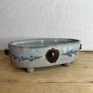May include: Oval ceramic planter with a speckled, light blue and brown glaze. The planter has three small feet and two side handles. A dark brown circular accent is on the front. The planter sits on a wooden surface.