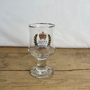 May include: Clear glass goblet with a silver rim. The goblet features a crown and laurel wreath design with the text "1977 THE QUEEN'S SILVER JUBILEE". The goblet has a round base and a stem.