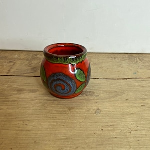 Puede incluir: Una pequeña maceta redonda de cerámica con un vibrante esmalte rojo. La maceta presenta un borde marrón oscuro y detalles verdes en la parte superior. Patrones decorativos de remolinos azules y verdes adornan el exterior. Esta maceta decorativa es adecuada para plantas pequeñas o como objeto decorativo.