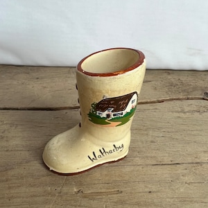 May include: Cream-colored ceramic boot-shaped vase with a painted cottage scene and the word "Wathby" in black script. The boot has a brown rim and laces, and sits on a wooden surface.