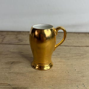 Puede incluir: Una pequeña taza dorada con asa. La taza tiene un acabado metálico brillante y un interior blanco. La base de la taza tiene un diseño acanalado. La taza está sobre una superficie de madera.